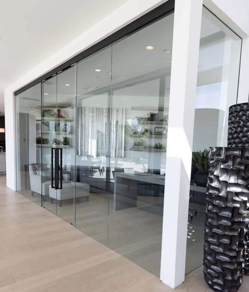 A modern home office enclosed in floor-to-ceiling glass walls with dark metal hardware, featuring a desk, plush seating, floating shelves, and light hardwood flooring.