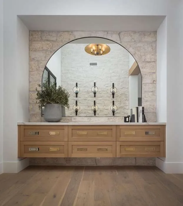 A floating light wood cabinet with brass hardware set inside a stone-backed wall nook, featuring a large arched mirror, a potted plant, and light hardwood flooring.