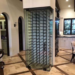 A modern glass-enclosed wine rack stands in a spacious, elegant kitchen with tiled floors, white cabinetry, and arched doorways. The wine rack is empty and features black metal holders inside.