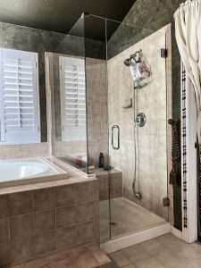 Modern Monterey bathroom in CA with a glass-enclosed shower beside a bathtub. The shower features a hand-held showerhead, soap dish, and beige tiles. White shutters dress the windows, complemented by a nearby white curtain.