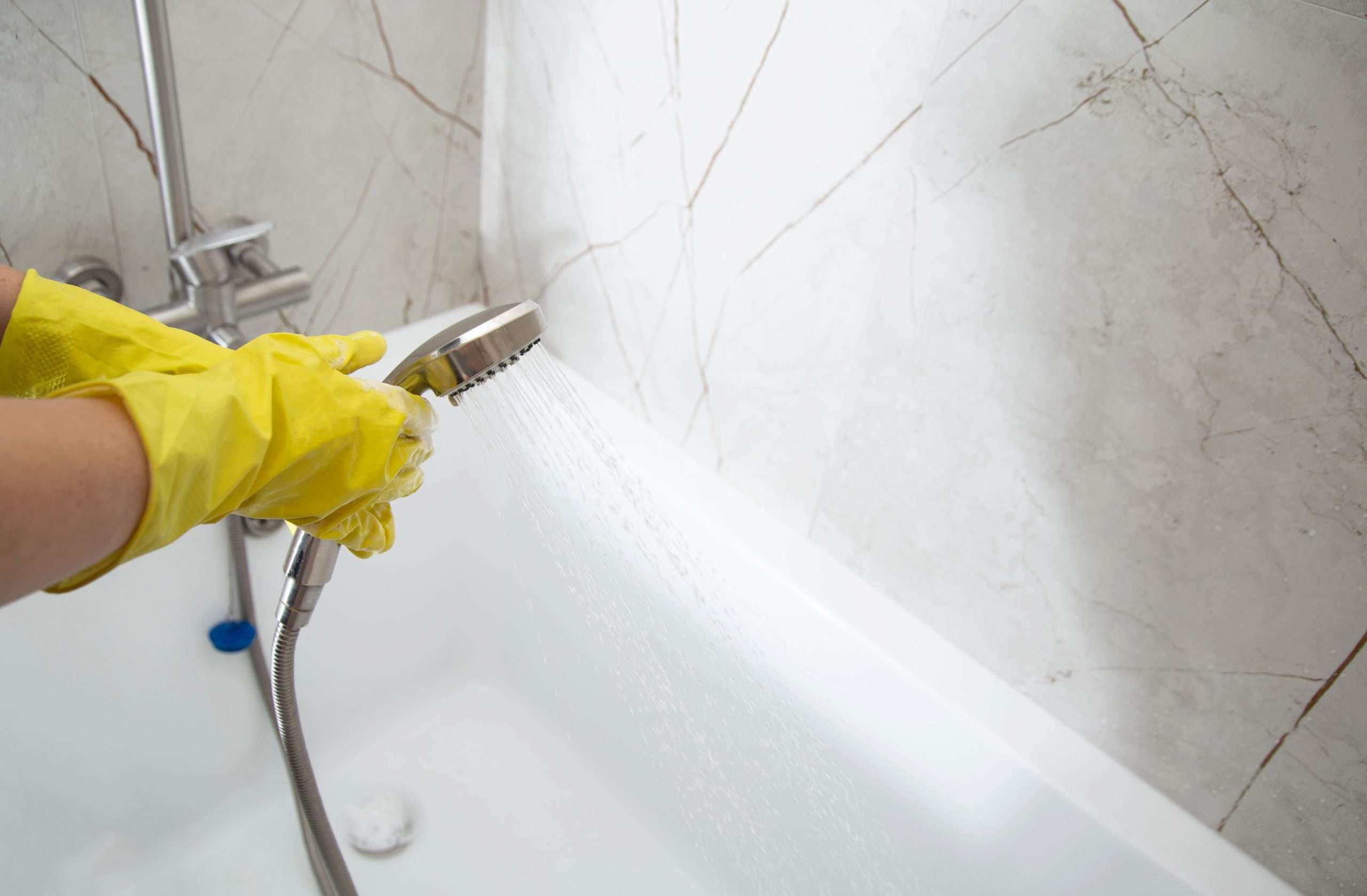 A hand wearing a yellow glove holds a showerhead, spraying water into a clean, white bathtub against a light marble wall.