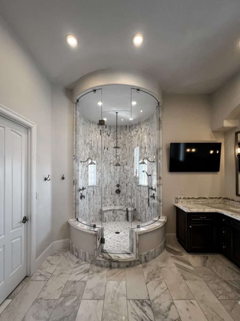 A luxurious, curved, all-glass shower stall with multiple shower heads and a built-in bench. The shower's interior and floor are covered in geometric marble mosaic tiles, while the exterior floor and base are large marble tiles. A vanity with a dark wood cabinet and a marble countertop is visible to the right.